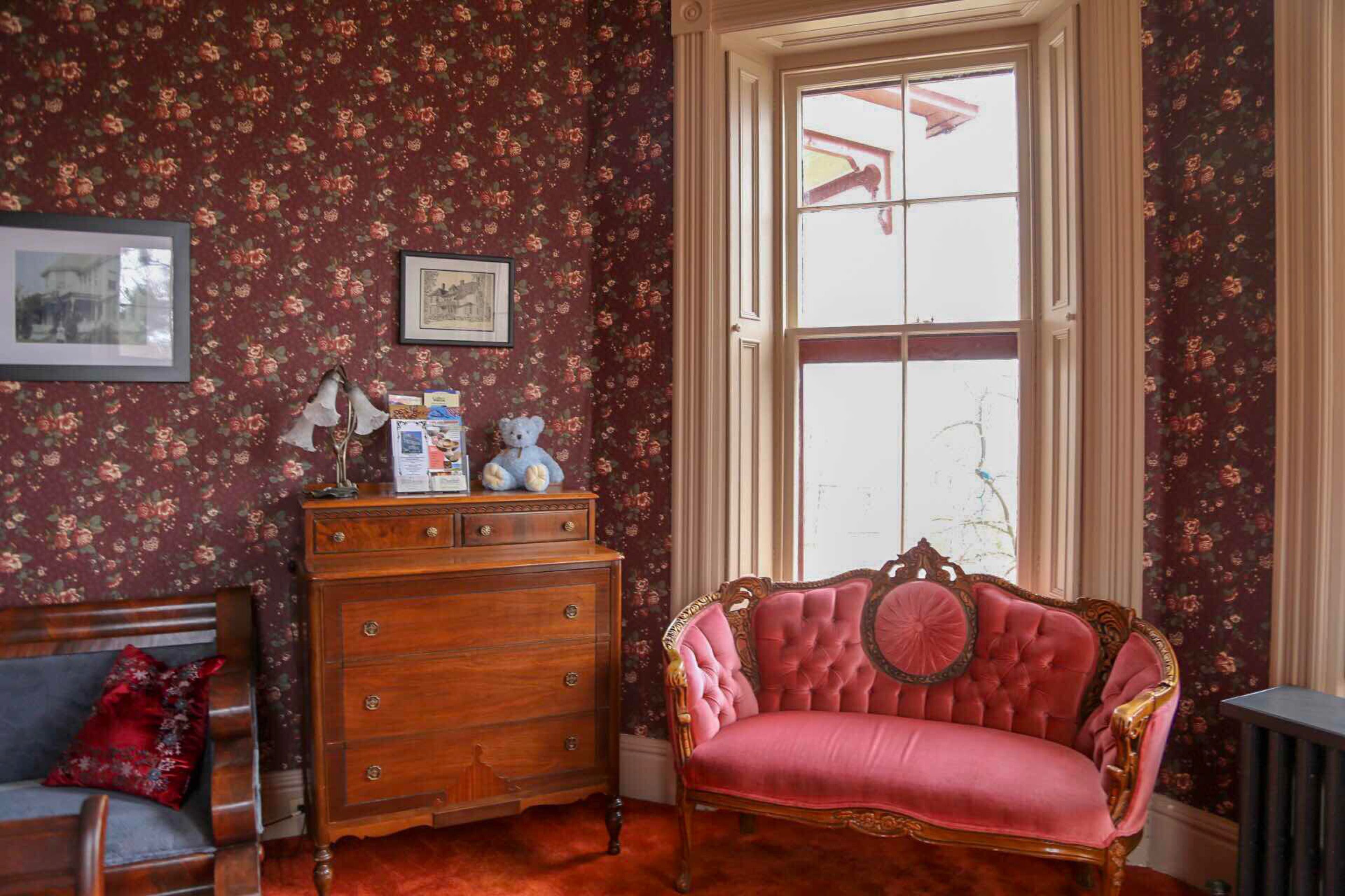 Una habitación con papel tapiz floral, un aparador de madera, una butaca tapizada en rosa y una ventana con vistas a una estructura roja.