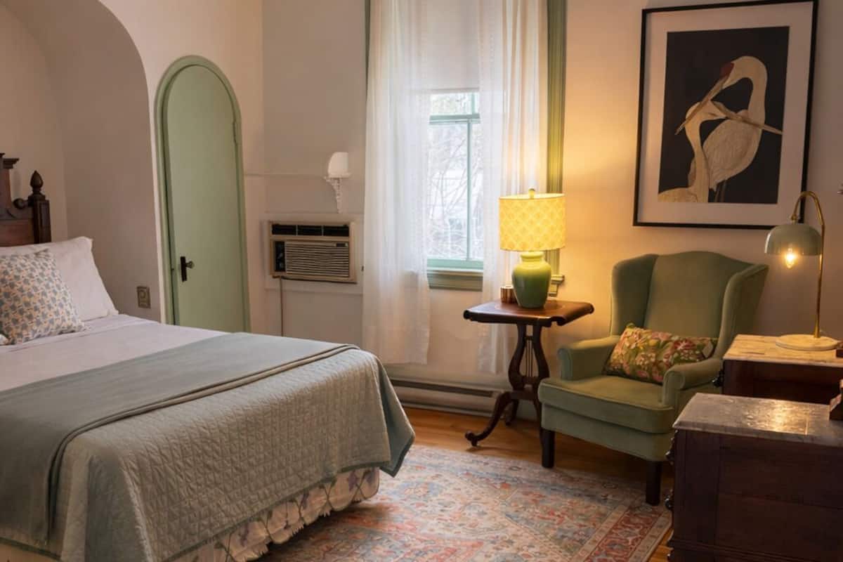 Une chambre à coucher avec un lit, une chaise, une lampe, une fenêtre avec des rideaux, un tableau encadré et une porte.