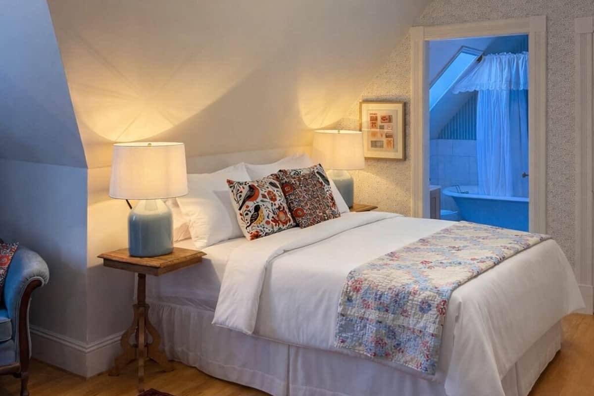 Une chambre avec un plafond en pente, un lit recouvert d’une literie blanche, un tapis à motifs et une petite table de chevet avec une lampe.