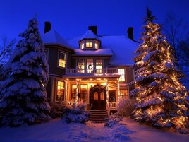 Una casa grande de dos pisos con un porche que rodea toda la estructura, árboles cubiertos de nieve y un árbol de Navidad iluminado.