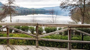 Une terrasse en bois donnant sur un lac paisible, avec une vue lointaine sur des bâtiments et des arbres.