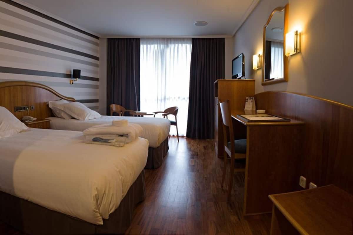 Een hotelkamer met twee bedden, een bureau, een stoel en een groot raam met gordijnen.