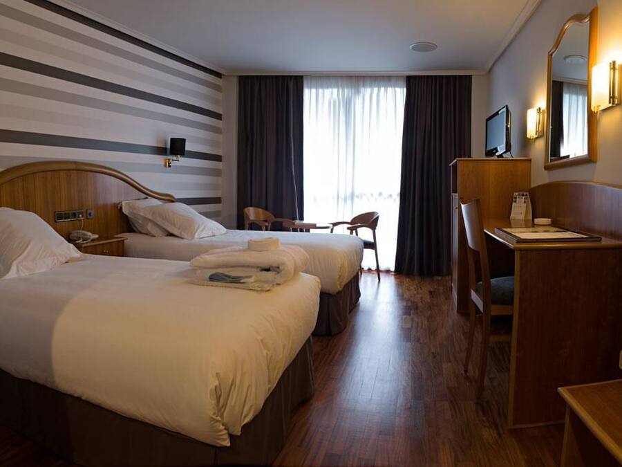 A hotel room with two beds, a desk, a chair, and a large window with curtains.
