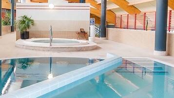 An indoor swimming pool with a hot tub, surrounded by plants and a tiled wall.