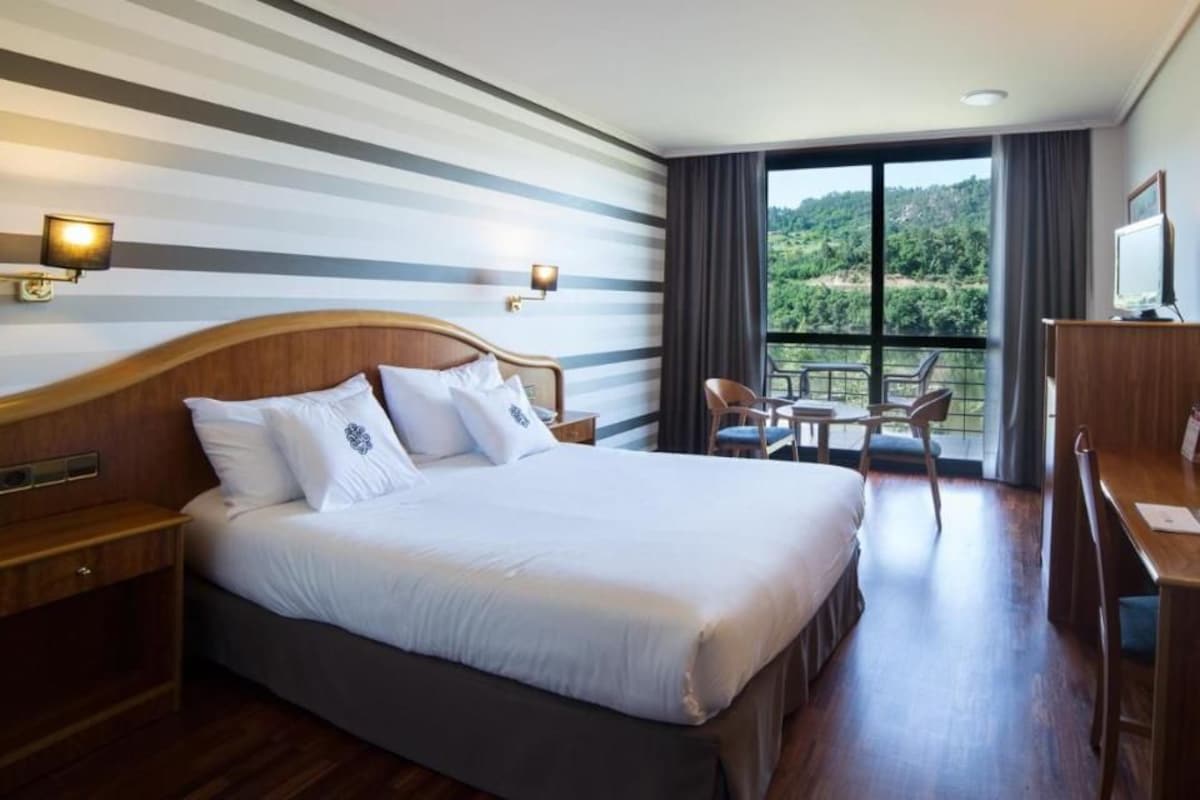 Habitación de hotel con una cama grande, un escritorio y un balcón con vistas a zonas verdes.
