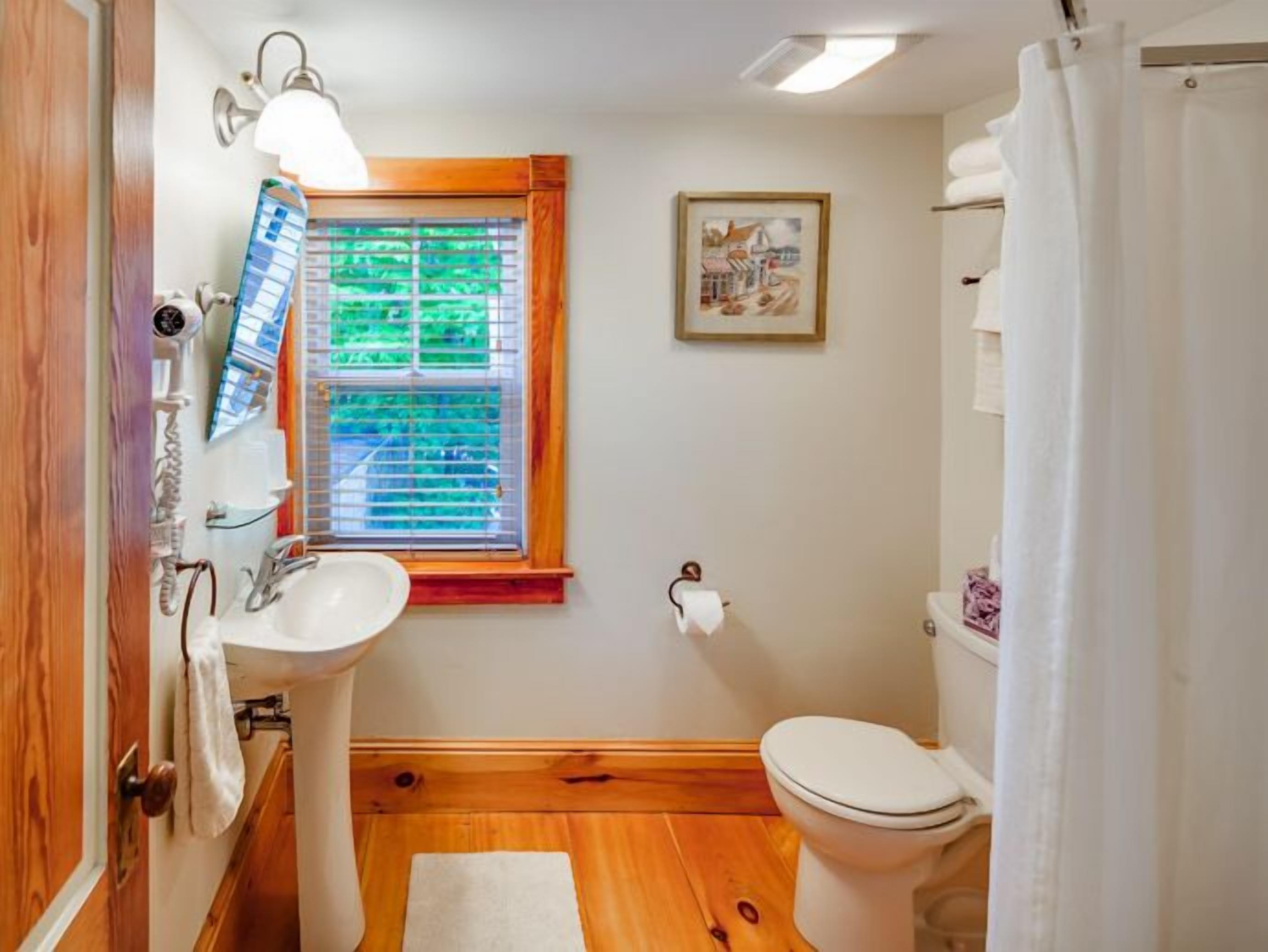 A bathroom with a wooden floor, a white sink, a toilet, a window with blinds, and a framed picture on the wall.