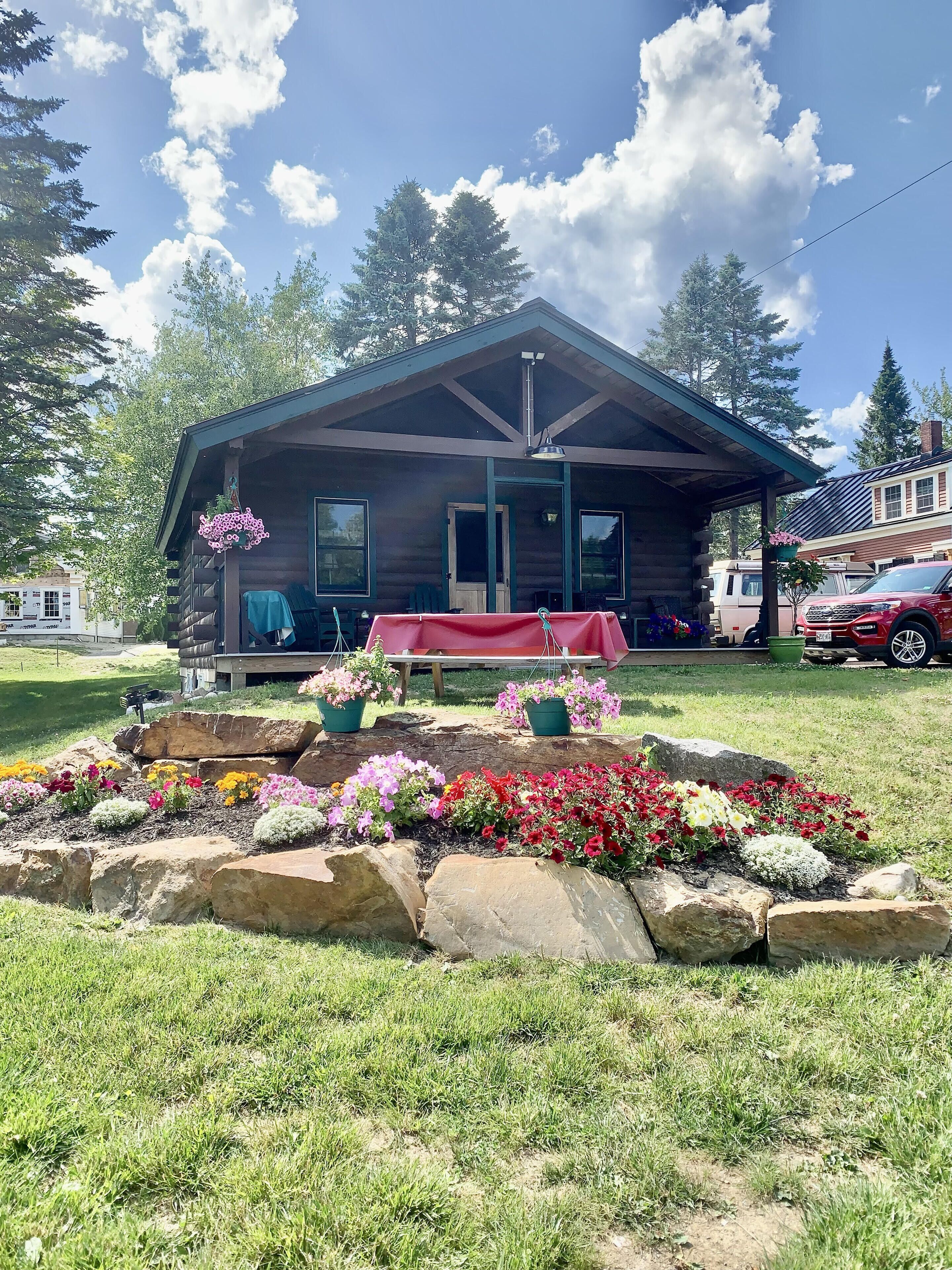 A log cabin with a porch, surrounded by a flower bed and a picnic table.