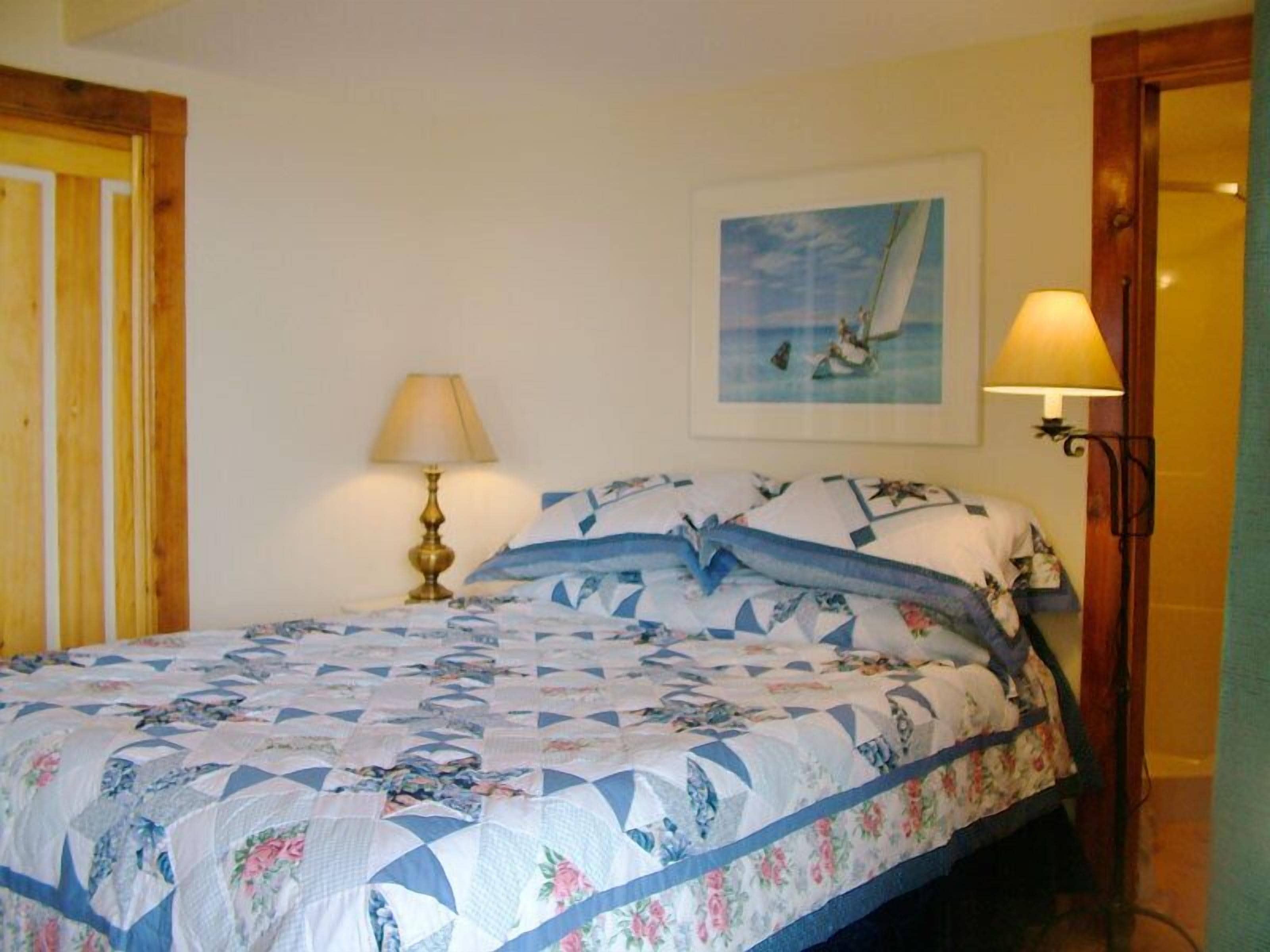 A bedroom with a bed covered in a floral quilt, a painting of a sailboat on the wall, and two wall-mounted lamps.