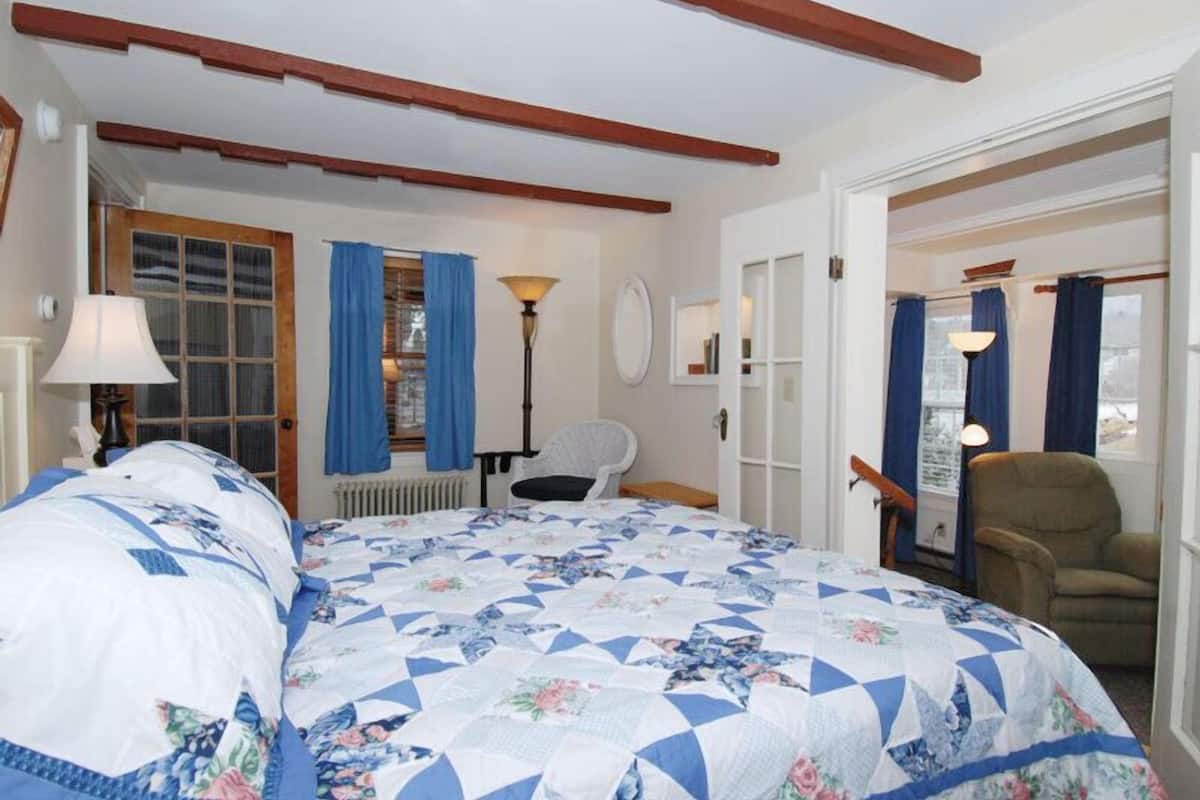 Ein Schlafzimmer mit einem großen Bett, einem Sessel, einer Lampe und einem Fenster mit Vorhängen.