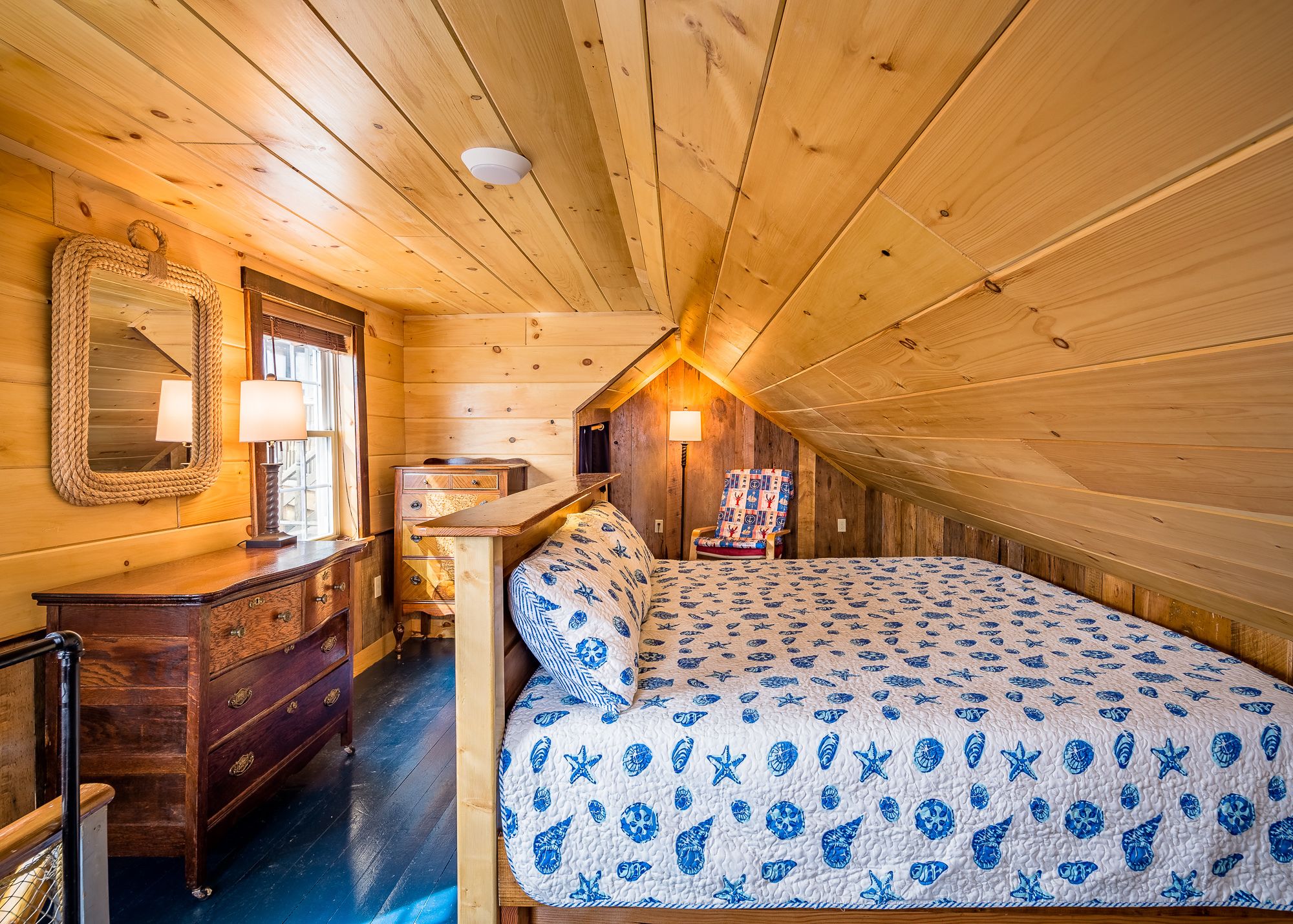 A cozy attic bedroom with a wooden ceiling, a bed with a blue and white patterned bedspread, a wooden dresser, a chair, and a mirror with a rope frame.
