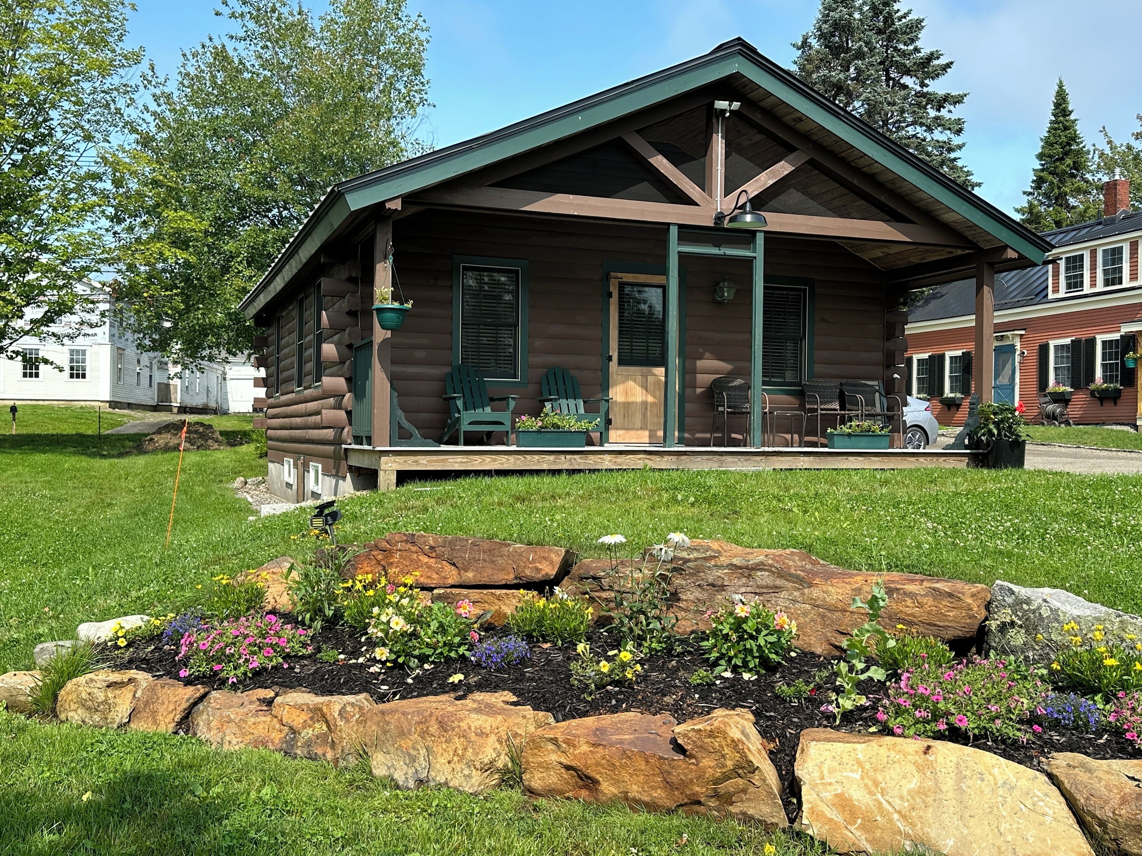 A log cabin with a porch, surrounded by a garden with various flowers and rocks.
