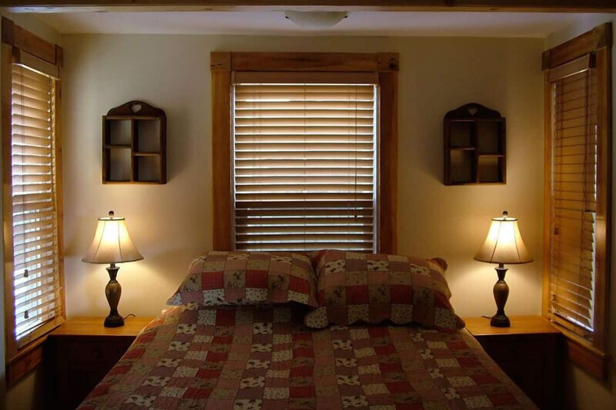 Ein Schlafzimmer mit einem Bett, zwei Nachttischen mit Lampen, einem Fenster mit Jalousien und Wandregalen.