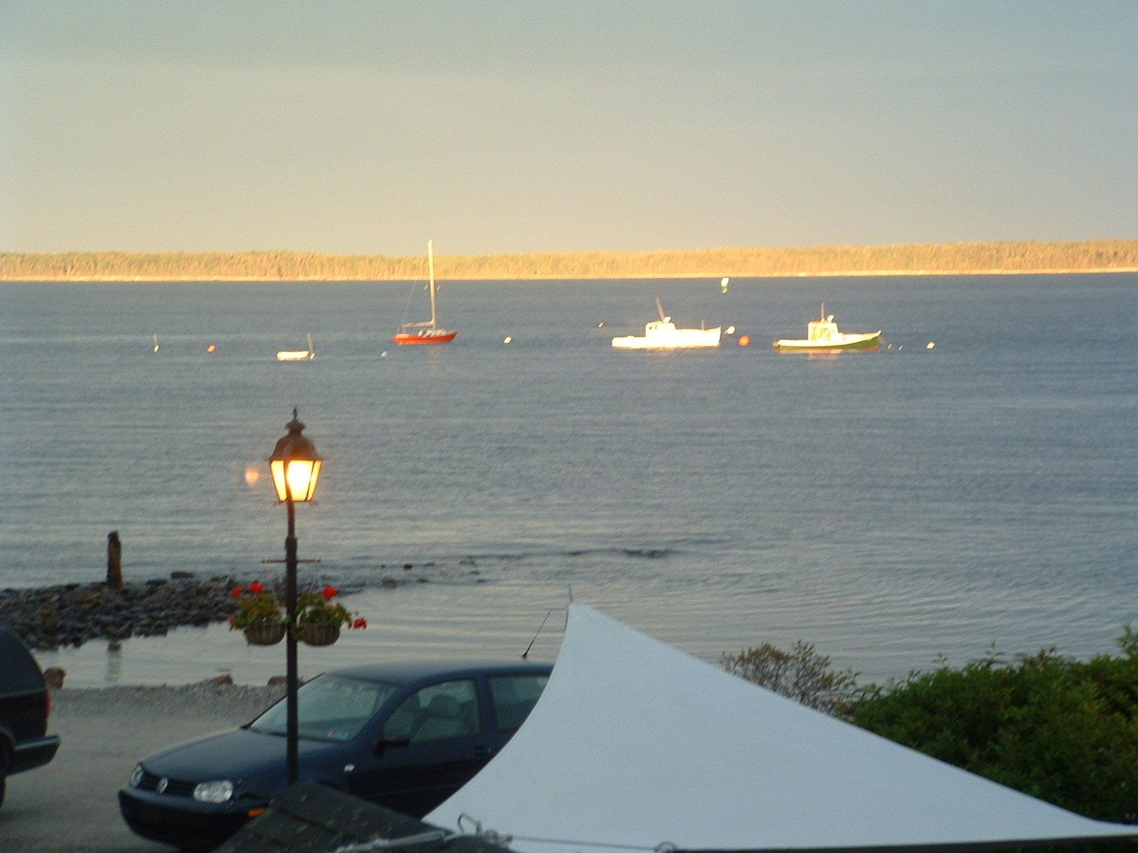 A coastal scene with a calm sea, a few boats, a street lamp, and a car parked near the shore.