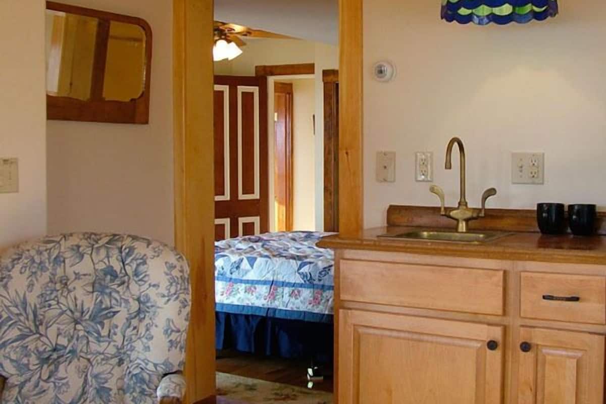 Ein gemütliches Zimmer mit Holzbalkendecke, einem stuhlförmigen Sitzmöbel mit Blumenmuster, einem Bett mit blau-weißer Steudecke und einer Küchenzeile mit Holzschränken und einem Spülbecken.