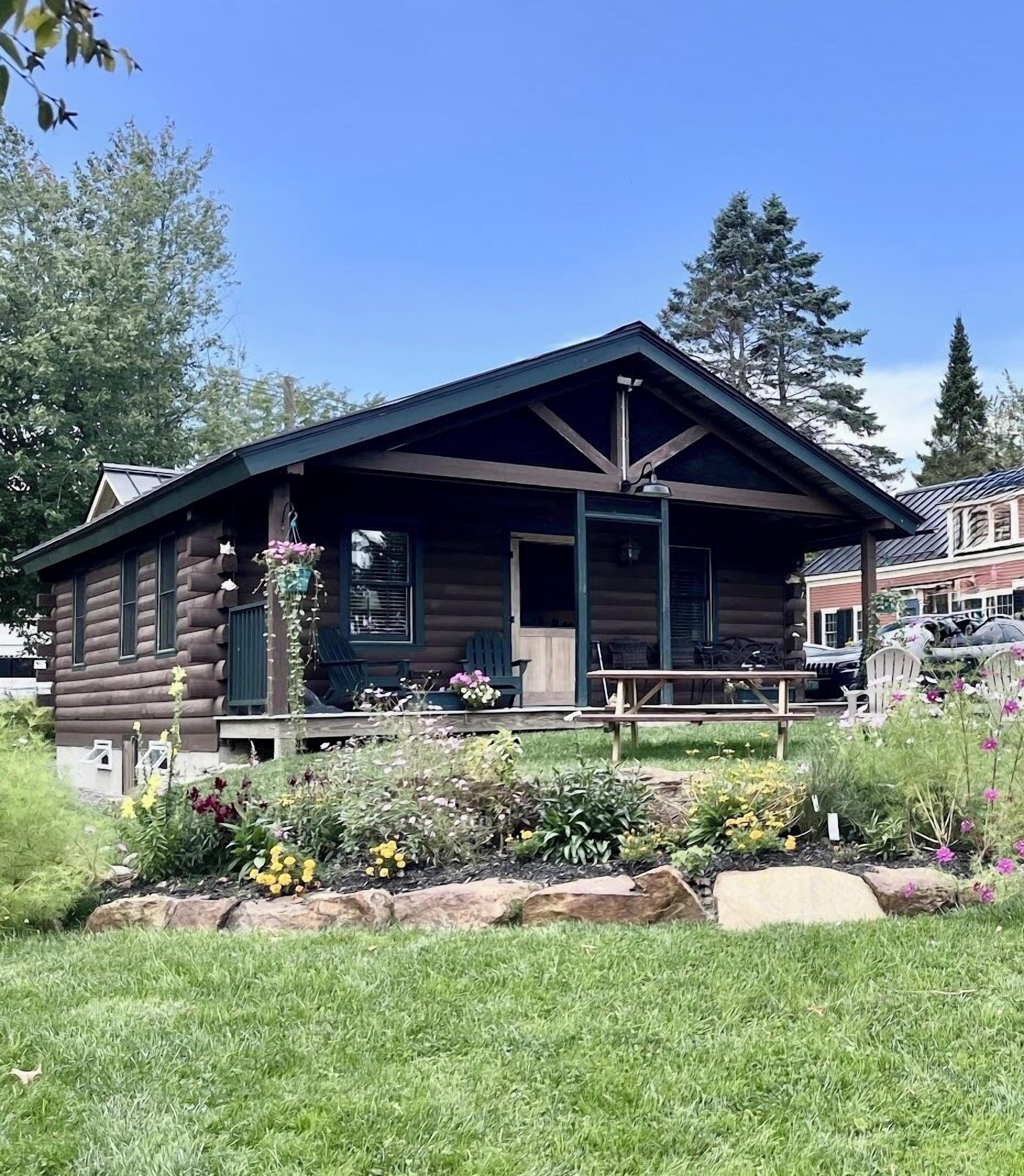 A log cabin with a porch, surrounded by a garden with various plants and flowers.