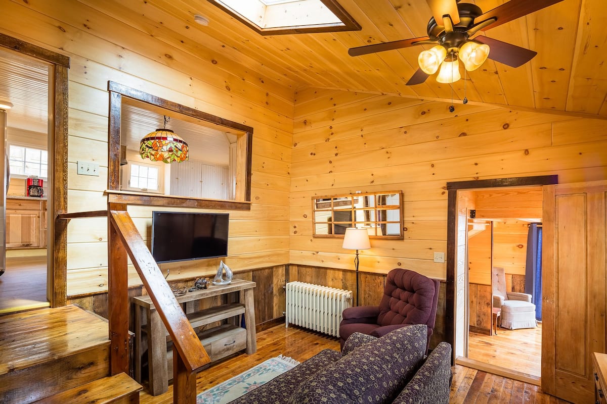 Ein gemütliches Blockhaushaus-Interieur mit Holzausstattung, Deckenventilator, Fernseher, Kamin und einer bequemen Sitzecke.