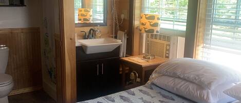 A bedroom with a bed, a window with blinds, a sink, and a mirror.