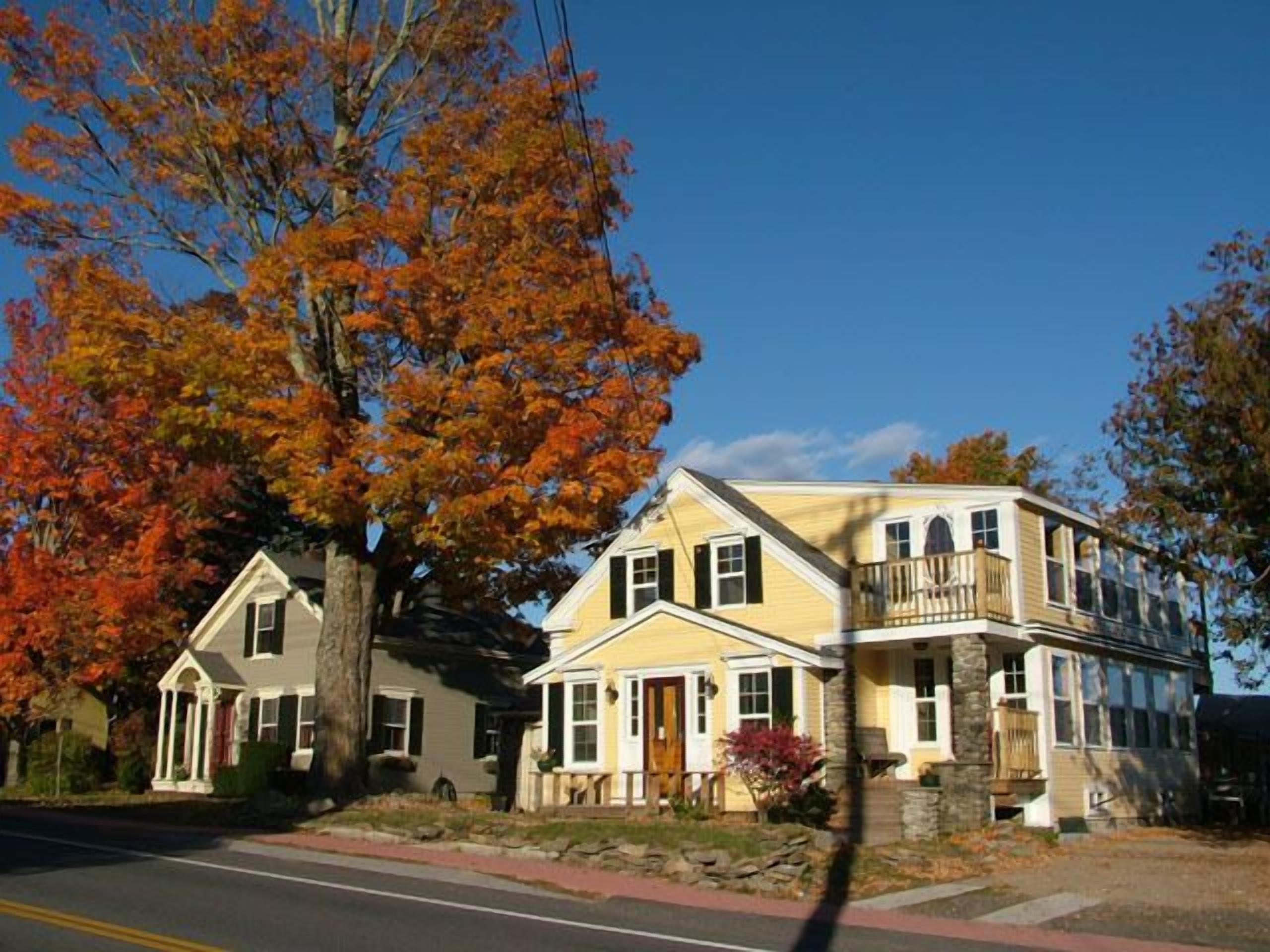 A two-story house with a front porch, a stone foundation, and a large tree with autumn leaves.