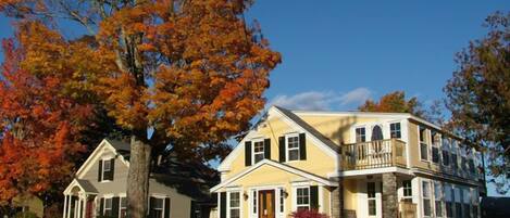 A two-story house with a front porch, a stone foundation, and a large tree with autumn leaves.