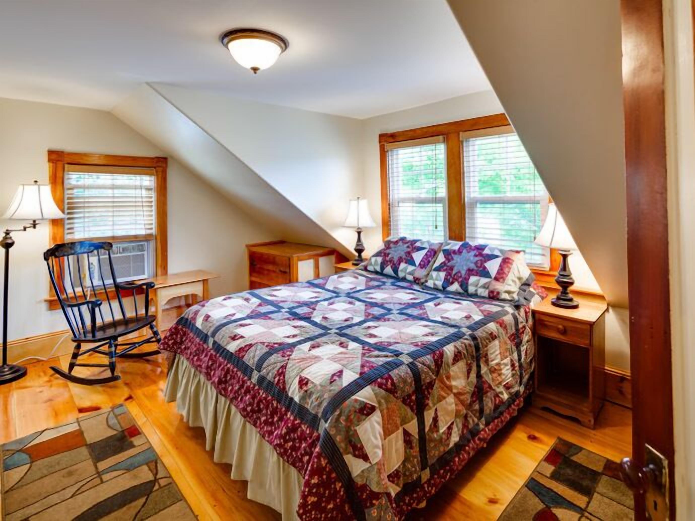 A bedroom with a large bed, two bedside tables with lamps, a rocking chair, and a window with blinds.