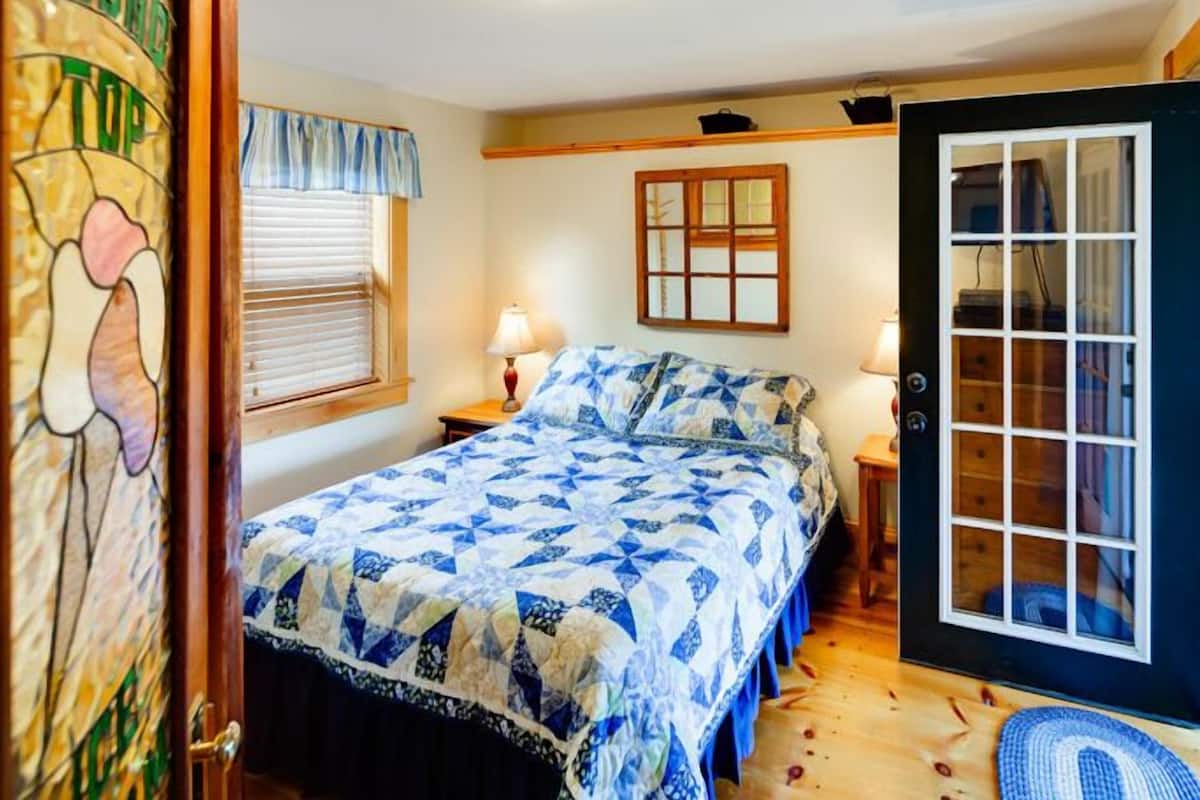 Ein Schlafzimmer mit einem Bett, zwei Nachttischen, einem Fenster mit Jalousien, einer Glastür und einer Wandleuchte.