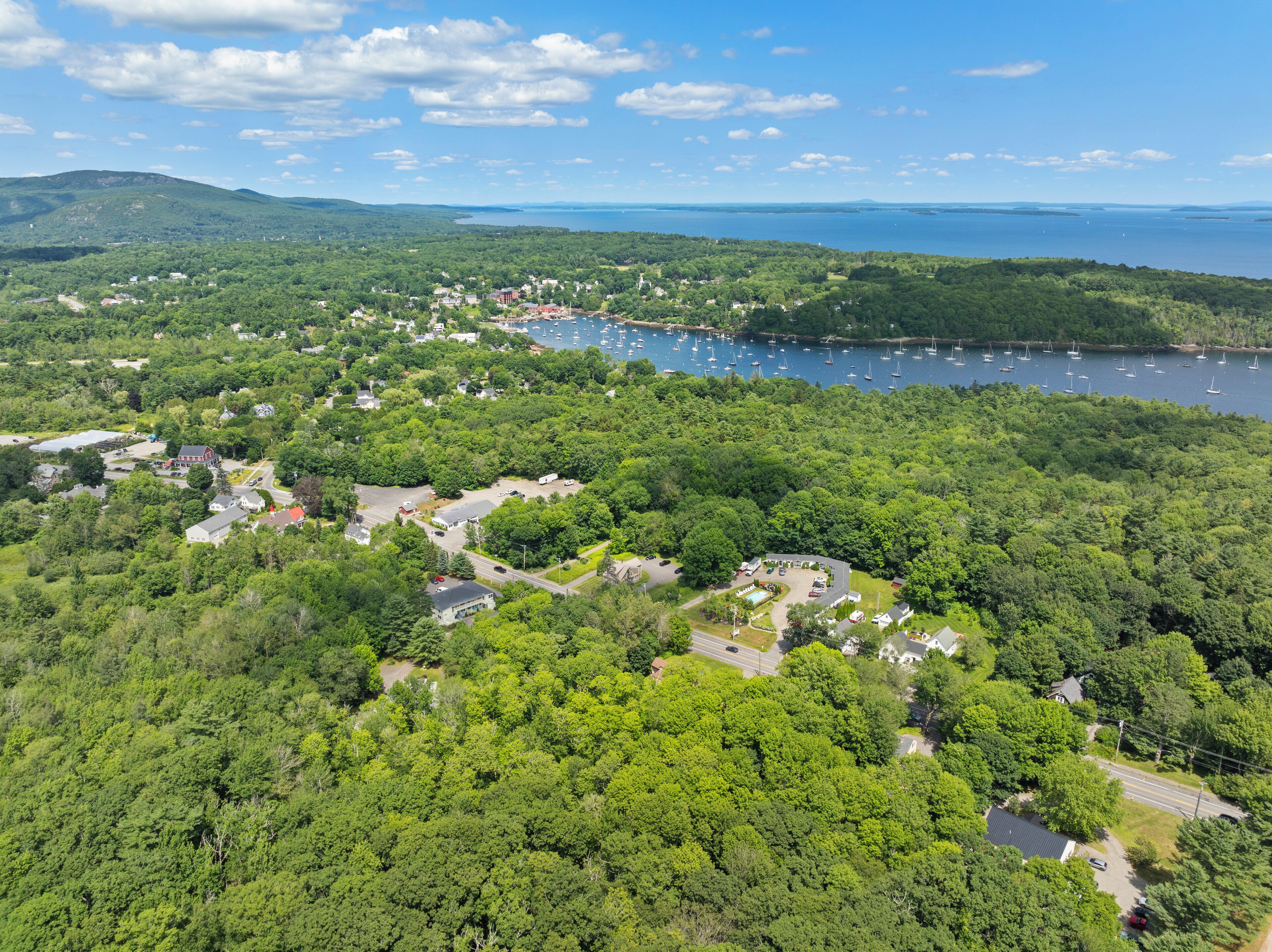 An aerial view of a coastal town with dense greenery, a marina with boats, and various buildings including a prominent red barn.