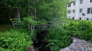 A wooden bridge over a stream in a lush garden with dense foliage and a white building in the background. - Harkness Brook Inn (Rockport)