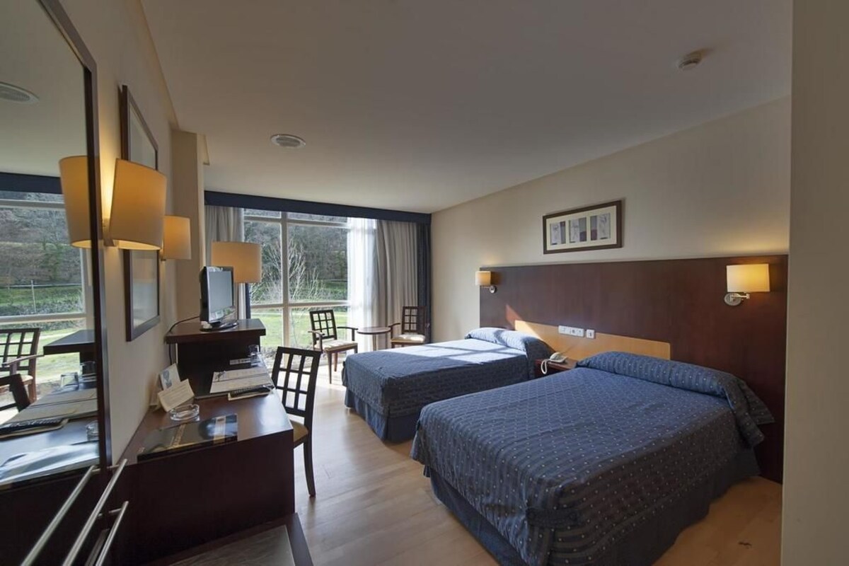 Ein Hotelzimmer mit zwei Betten, einem Schreibtisch mit Stuhl, einem Fernseher und einem Fenster mit Blick auf Grünflächen.