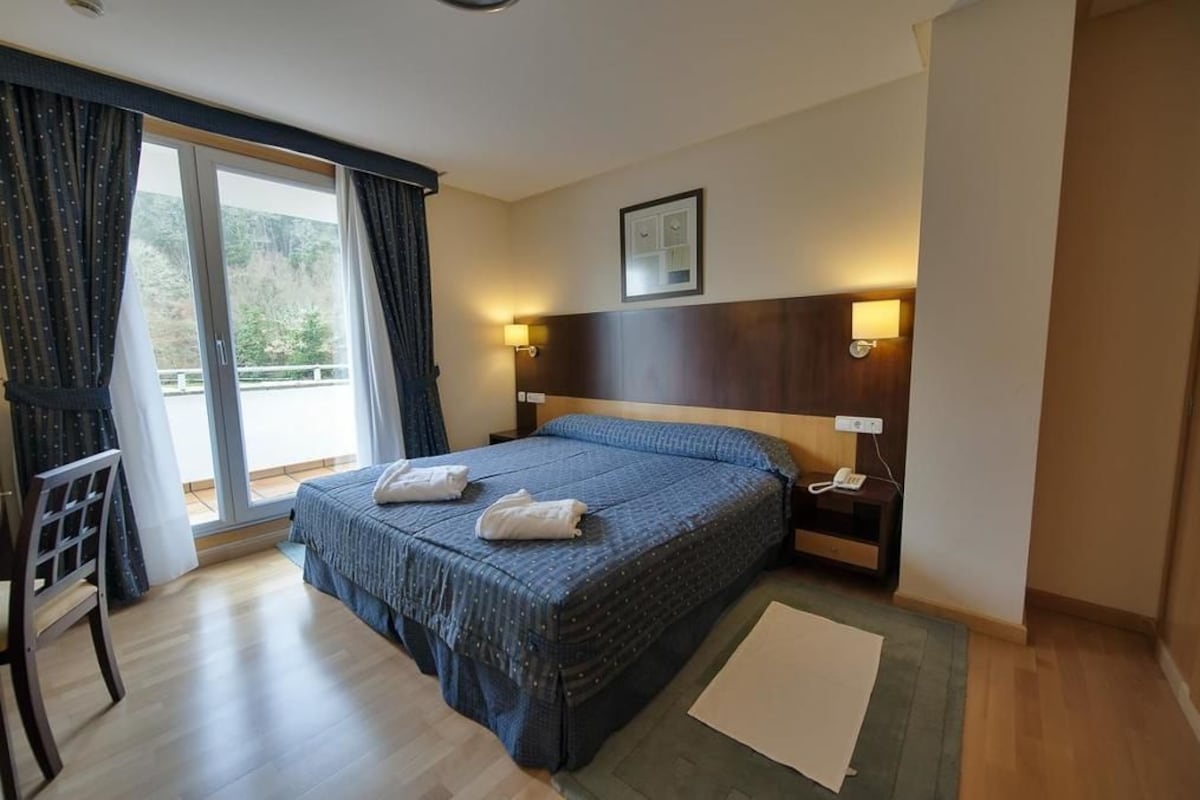 Hotelkamer met een groot bed, een bureau, een stoel, een nachtkastje met een lamp en uitzicht op de omgeving via een schuifdeur.