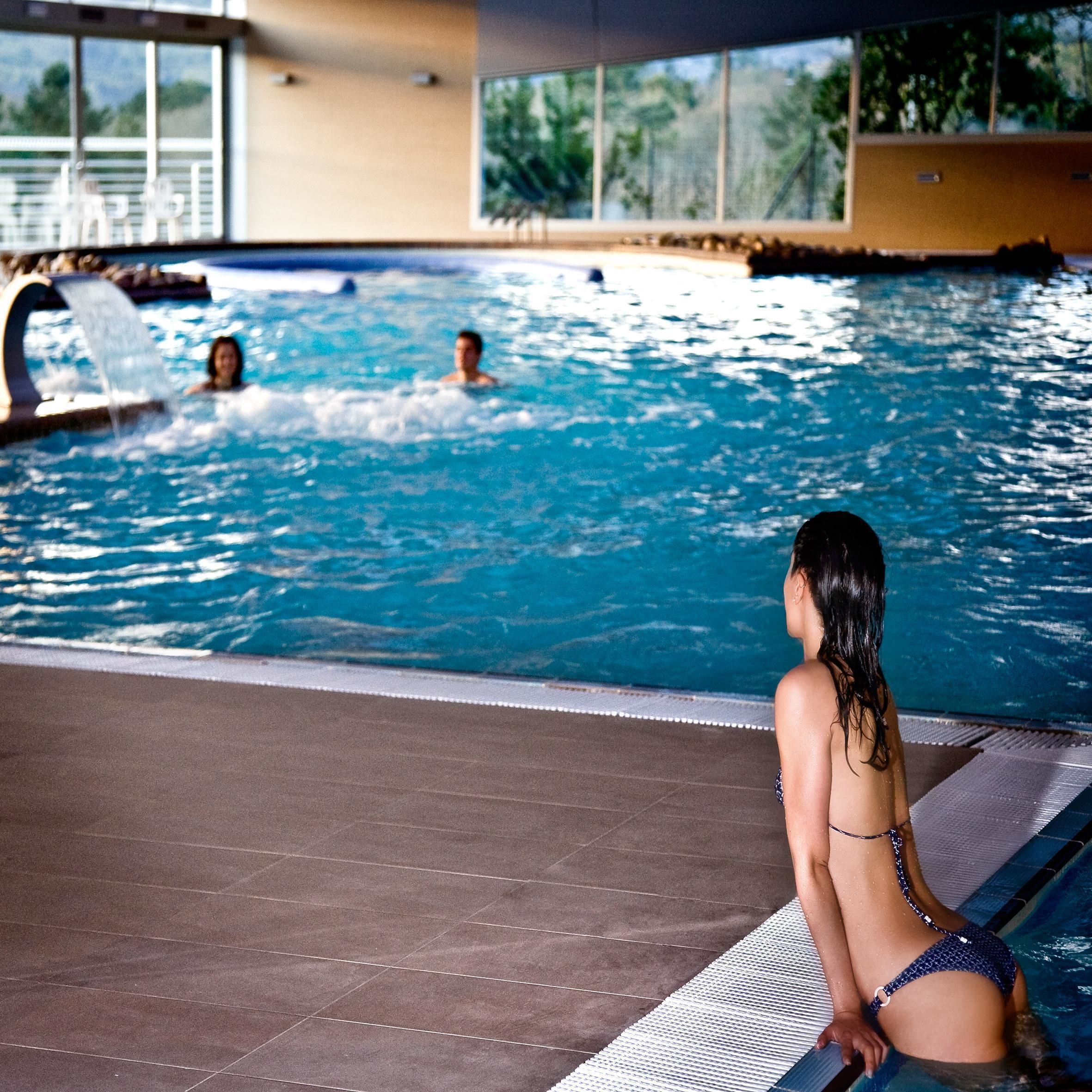 Una piscina interna con acqua limpida, pavimento piastrellato e una persona seduta sul bordo.