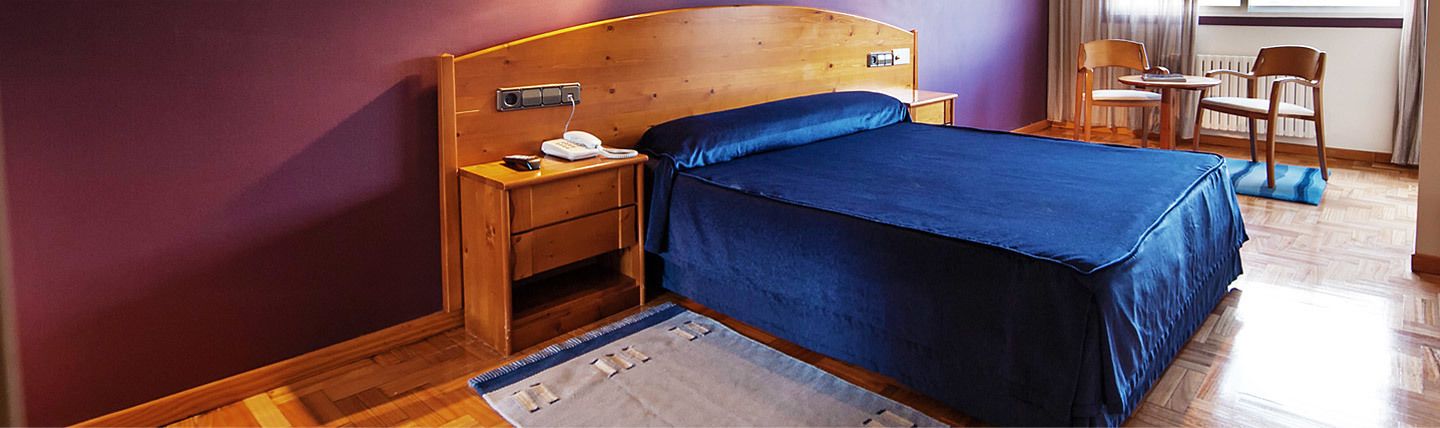A bedroom with a wooden headboard, a blue bedspread, a bedside table with a phone, and a chair near a window.