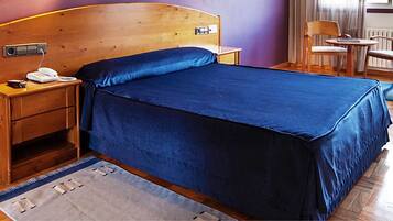 A bedroom with a wooden headboard, a blue bedspread, a bedside table with a phone, and a chair near a window.