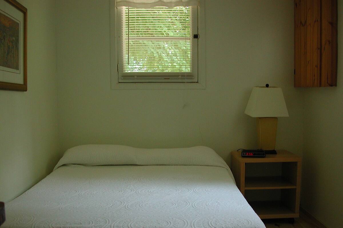 A single bed with a white bedspread in a room with a window, a nightstand with a lamp, and a wooden cabinet.
