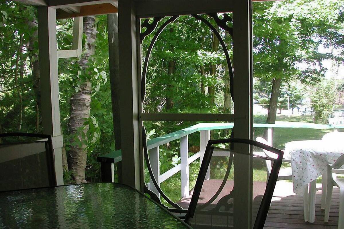 A covered outdoor seating area with a table and chairs, surrounded by greenery and a white railing.