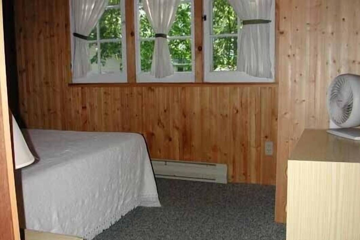 A small, single-room cabin with a bed, a window with curtains, a fan, and a wooden wall.