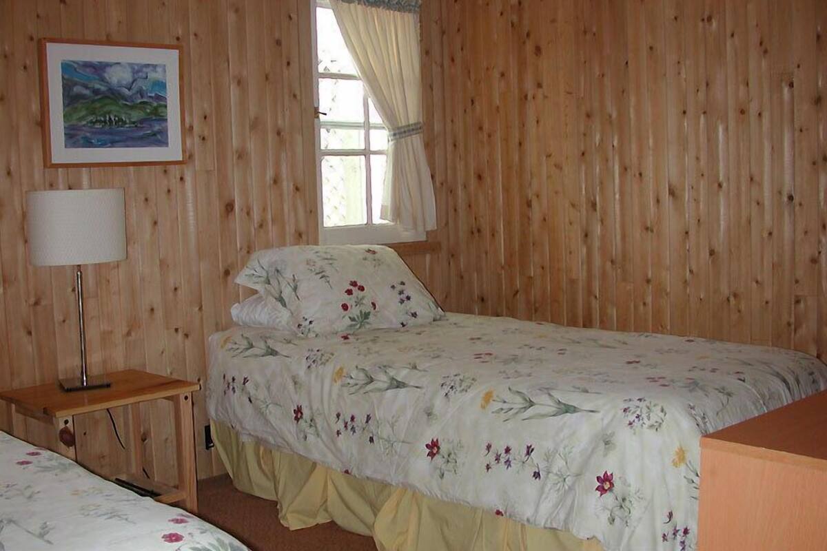 A room with wooden paneling, a bed with floral bedding, a bedside table with a lamp, and a window with curtains.