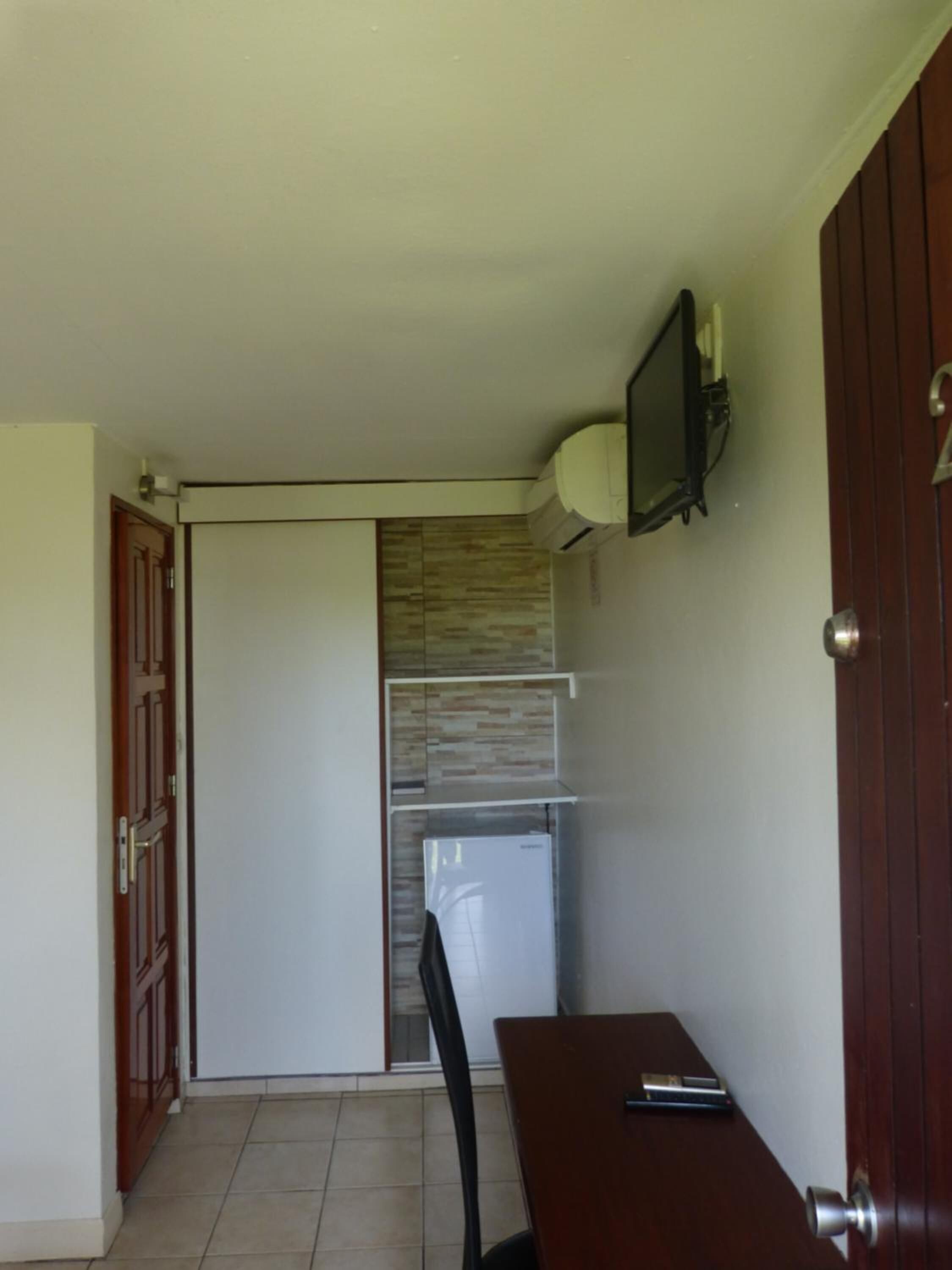 Habitación pequeña con puerta de madera, una mesita con silla, refrigerador blanco y televisor montado.