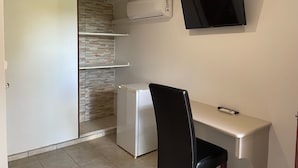 Een compacte kamer met een wit bureau, een zwarte stoel, een kleine witte koelkast, een wandtelevisie en een schuifdeurkast.