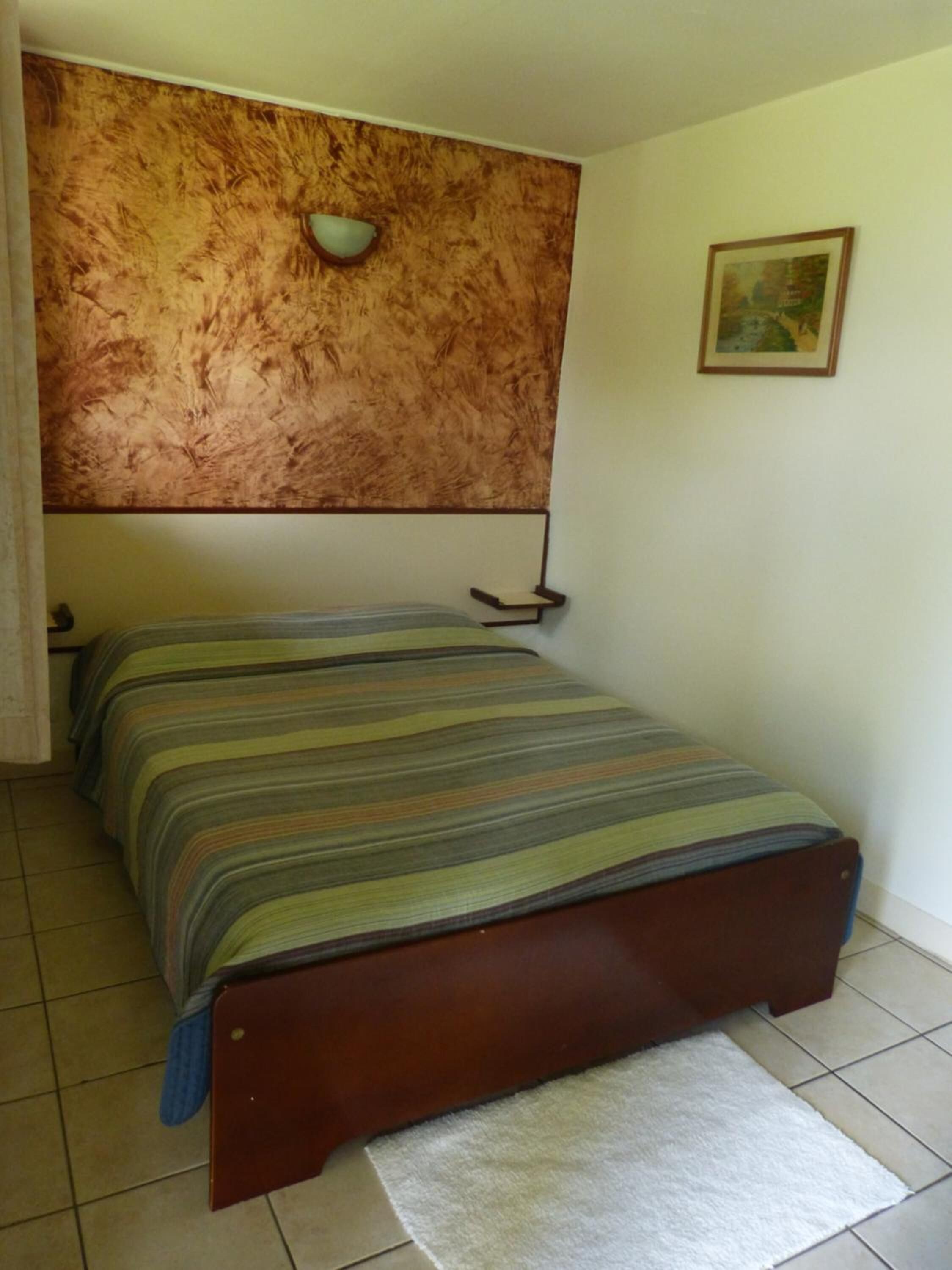 Une chambre à coucher comprenant un lit, une tête de lit, une table de chevet, une lampe fixée au mur et un tableau encadré.