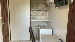 Een kleine, moderne kamer met een wit bureau, een zwarte stoel, een aan de muur bevestigde televisie en een betegelde vloer.