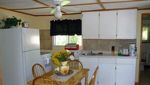 Une cuisine de taille modeste avec des meubles blancs, une table à manger en bois avec des chaises, un ventilateur de plafond et une fenêtre agrémentée de rideaux noirs.