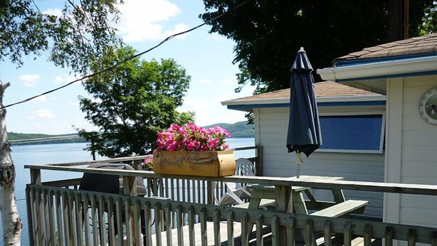 Une terrasse en bois avec une rambarde, une table et une chaise. On y trouve également un jardinier avec des fleurs roses et un parasol bleu.