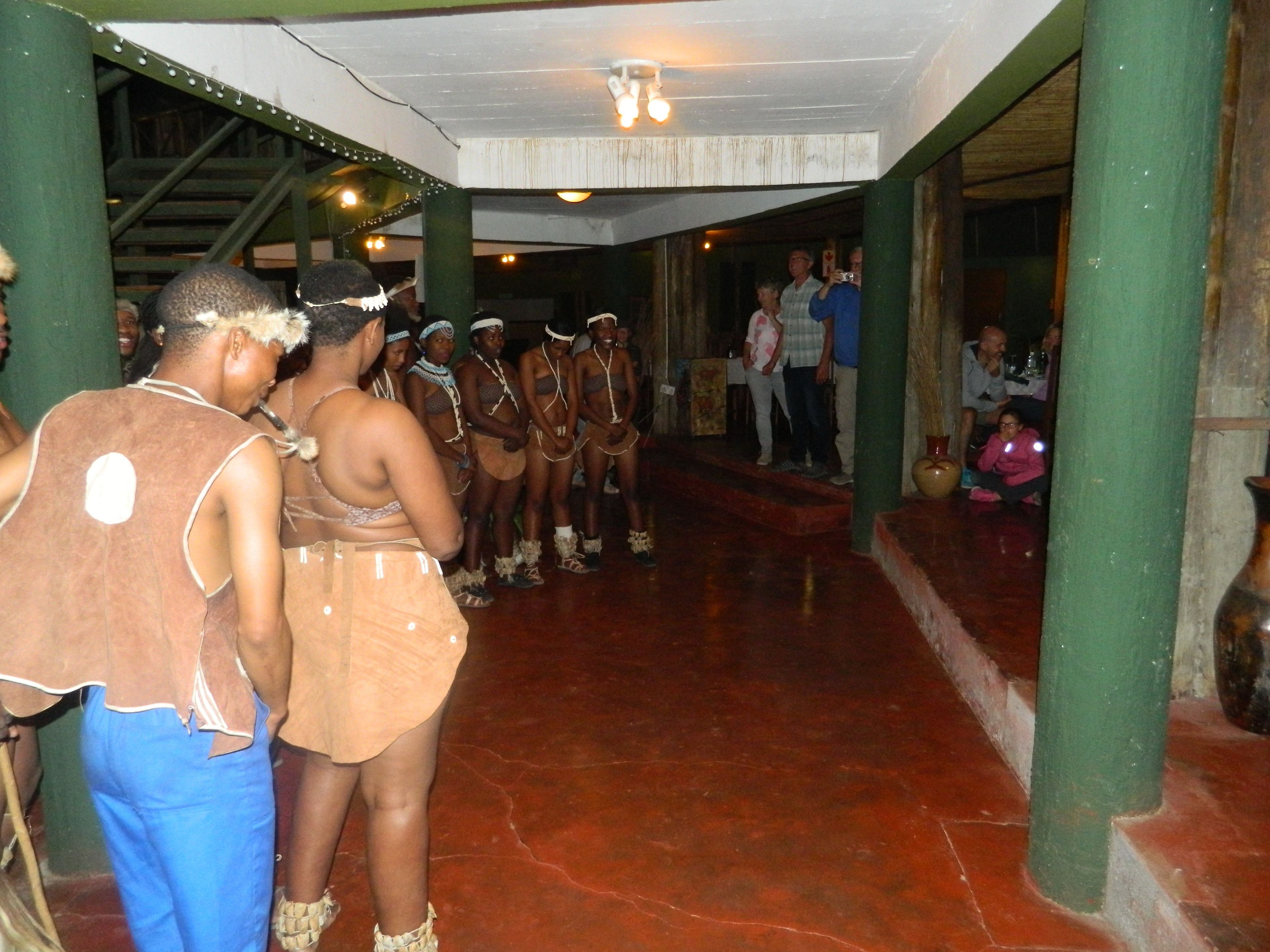 Un grupo de personas vestidas con atuendo tradicional se encuentran en un espacio cubierto con suelo de baldosas y pilares verdes.
