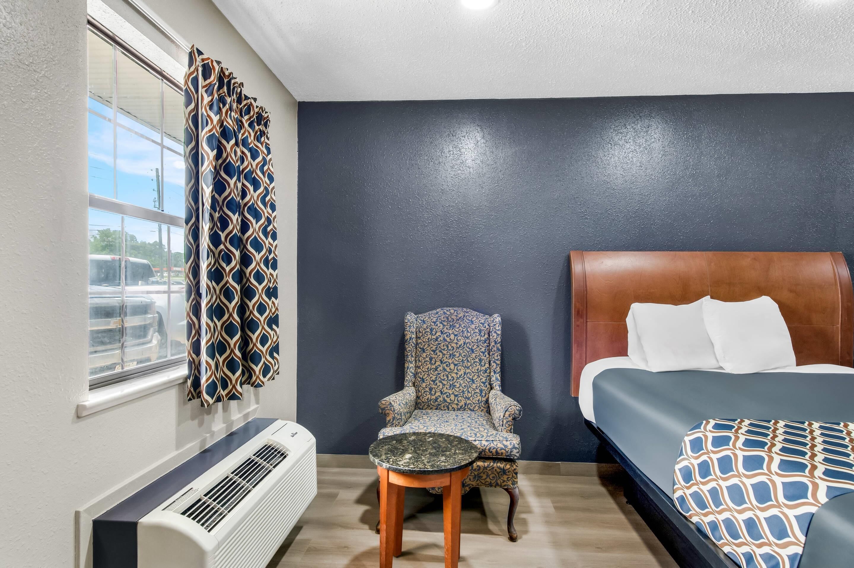 A hotel room with a bed, a chair, a table, a window with curtains, and a wall heater.