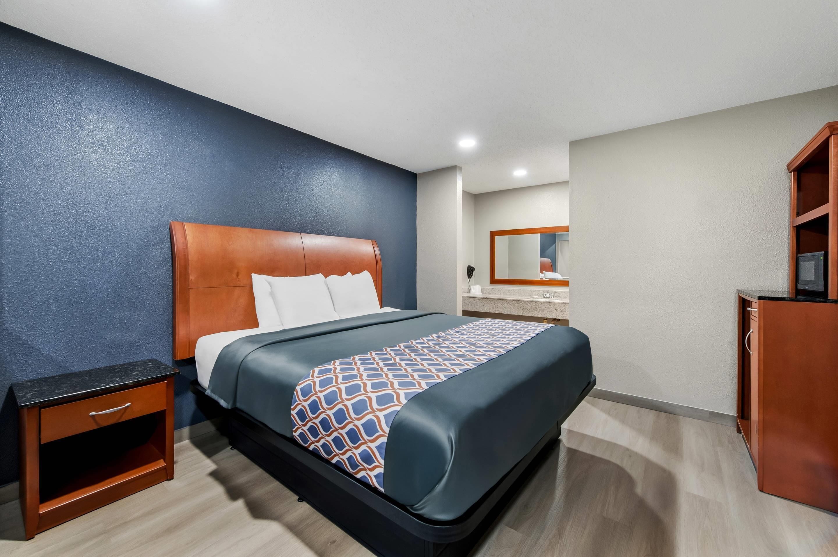 Habitación de hotel con una cama, una mesita de noche de madera, un aparador, un espejo y un baño a la vista.