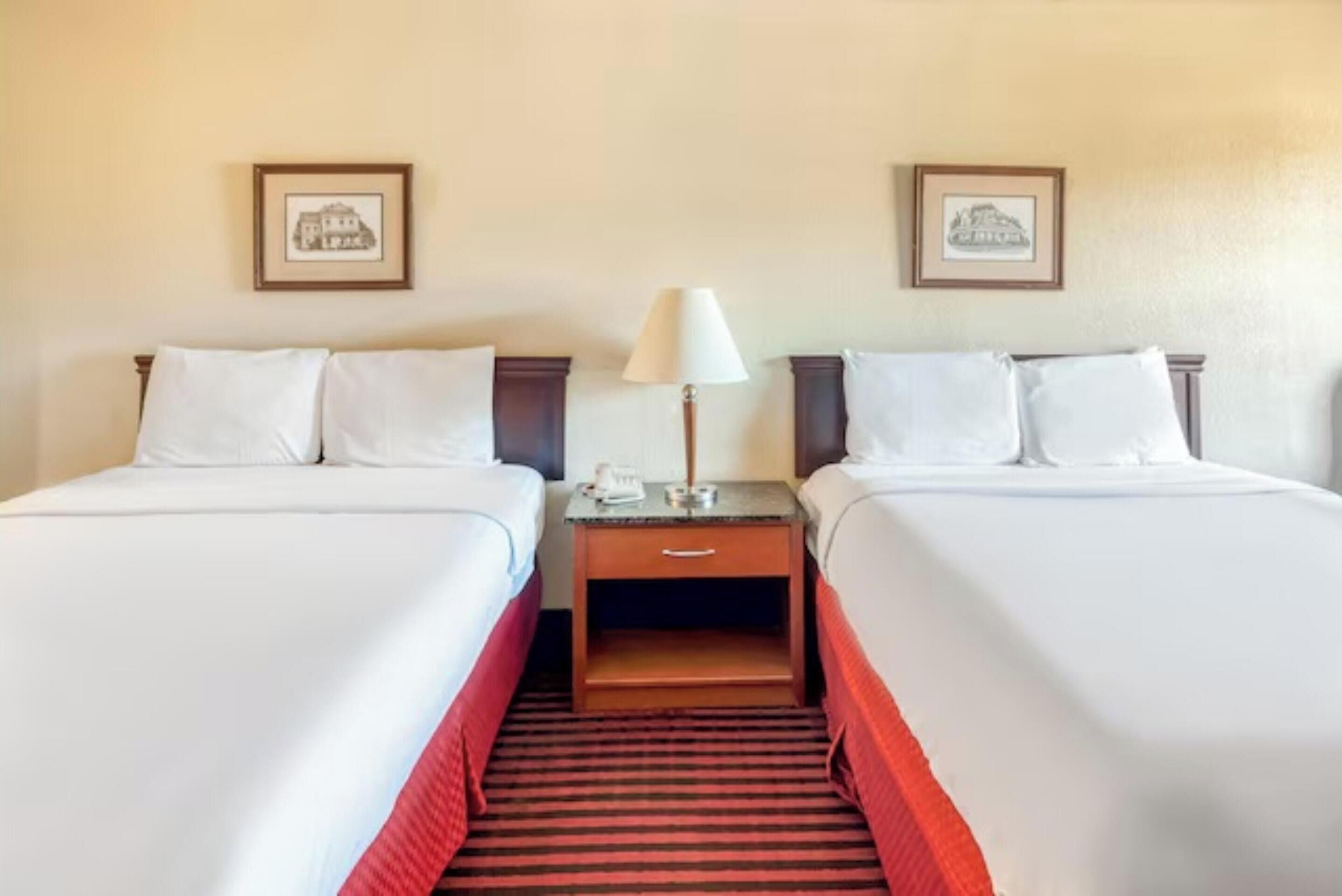 A hotel room with two beds, a nightstand with a lamp, and framed pictures on the wall.