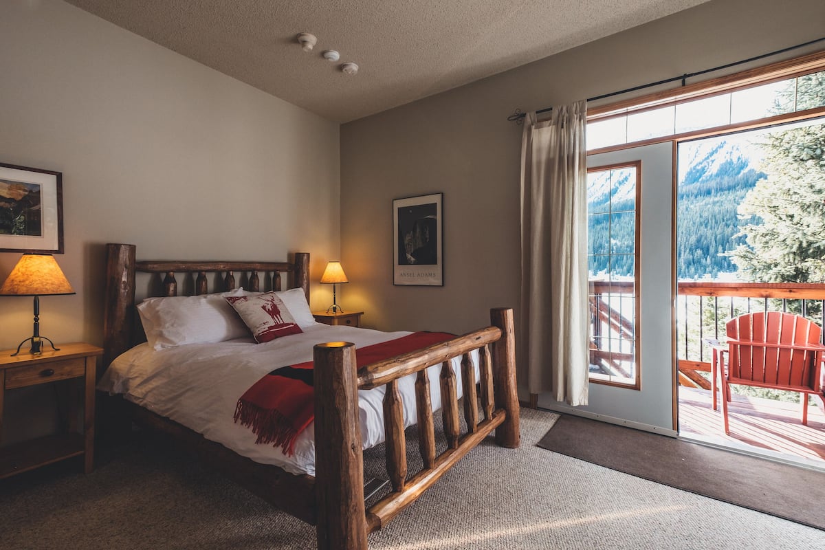 A bedroom with a wooden bed, a nightstand, a lamp, a framed picture, and a view of a balcony with a chair.