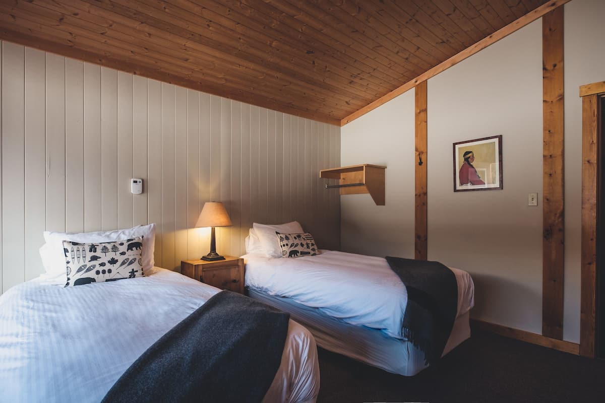 A room with two beds, wooden beams on the ceiling, a bedside table with a lamp, and a framed picture on the wall.