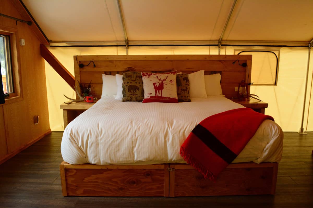 A wooden bed with a white mattress and red blanket, a wooden headboard, and a window on the left.