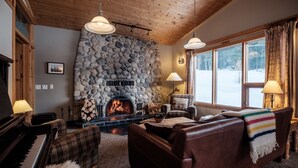 Una sala de estar acogedora con chimenea de piedra, un piano y un amplio ventanal con vistas nevadas.